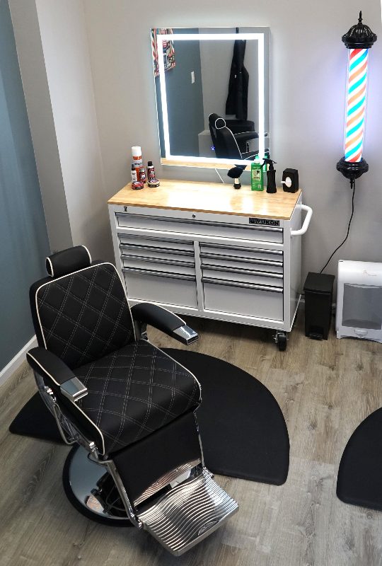A barber chair beside a lit mirror, tool chest and barber pole